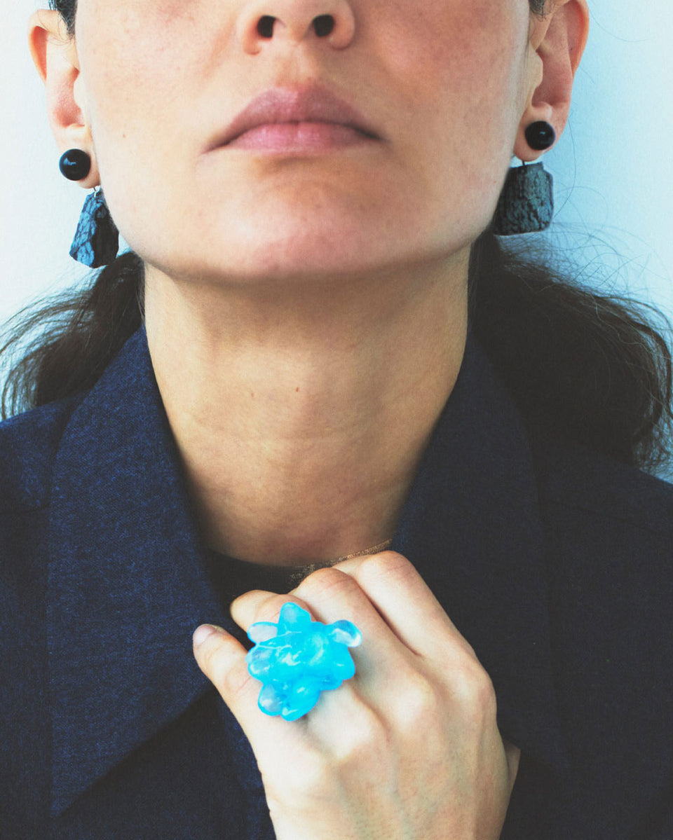Person wearing a blue flower-shaped ring with earrings and a dark outfit.