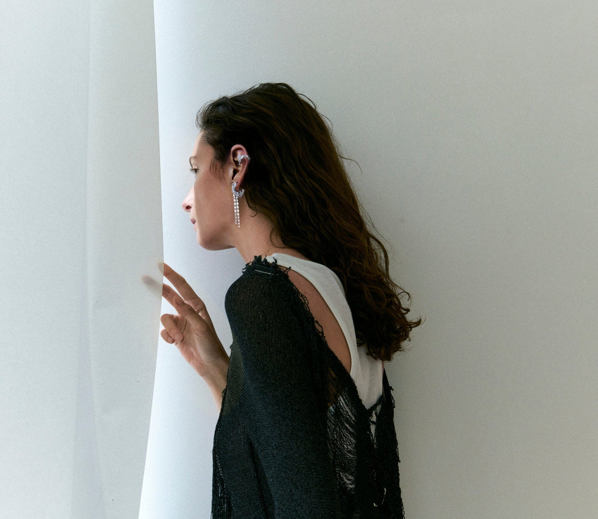 A woman in a black lace top and white undershirt stands in profile against a white wall, wearing a long silver dangle earring.