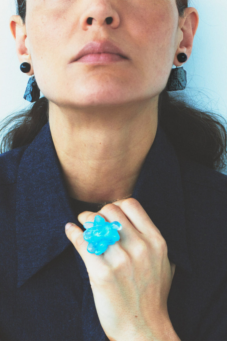 Person wearing a blue flower-shaped ring with earrings and a dark outfit.